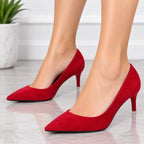 A pair of red pointed-toe high heels worn on a model's feet, shown against a clean white background with a marble floor and a potted plant.