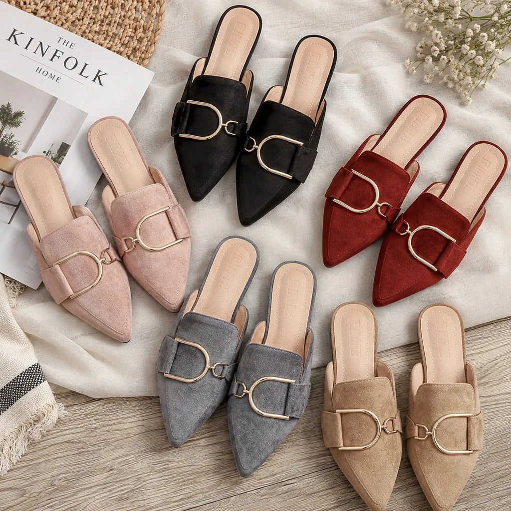 Six pairs of pointed-toe mule flats arranged on a light fabric surface over a wood floor. The flats are shown in blush pink, black, red, gray, and beige suede-like finishes, each with a large horsebit-style metal buckle on the vamp.