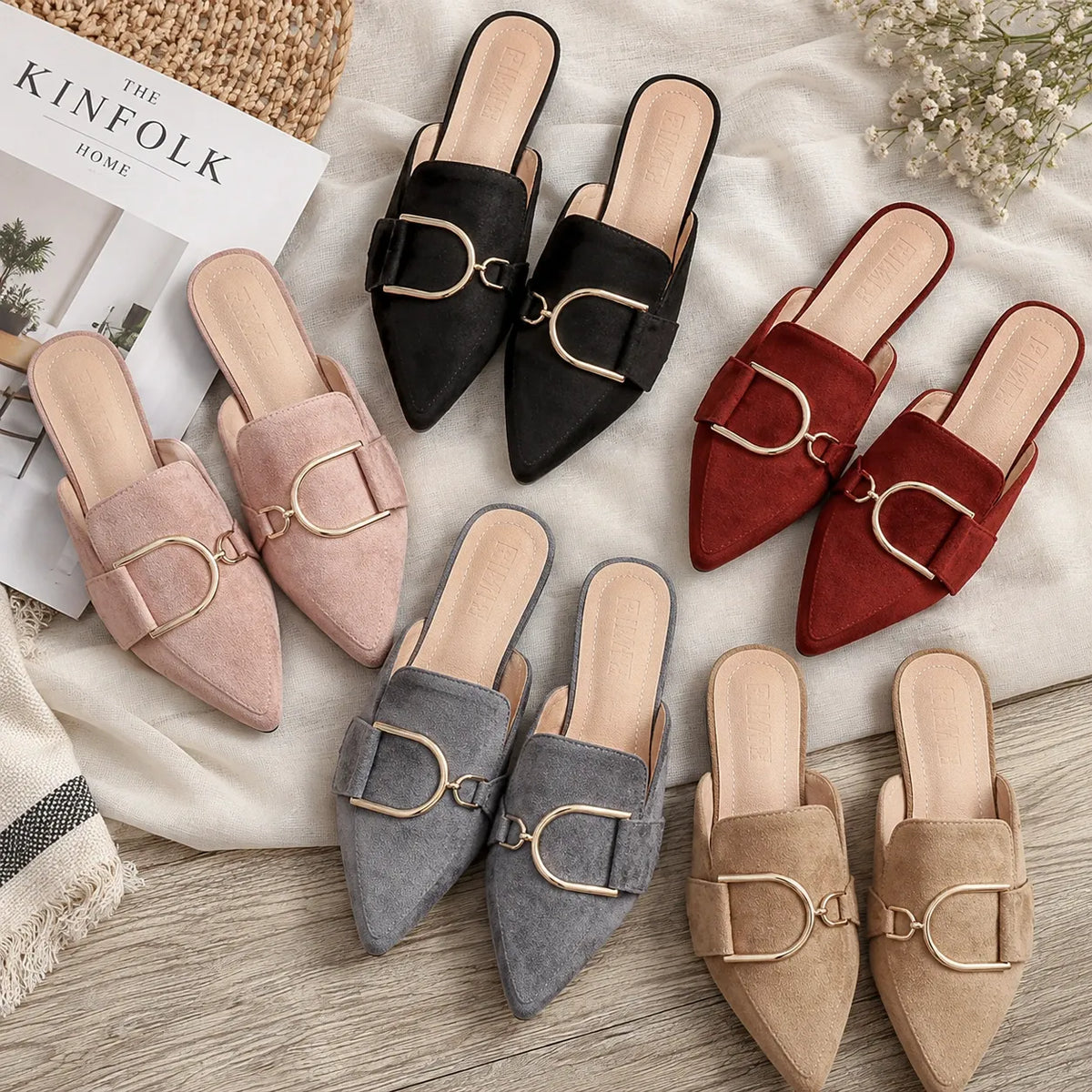 Six pairs of pointed-toe mule flats arranged on a light fabric surface over a wood floor. The flats are shown in blush pink, black, red, gray, and beige suede-like finishes, each with a large horsebit-style metal buckle on the vamp.