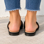 Black slide sandals with a low block heel and open toe, shown from the back on feet wearing frayed blue jeans.