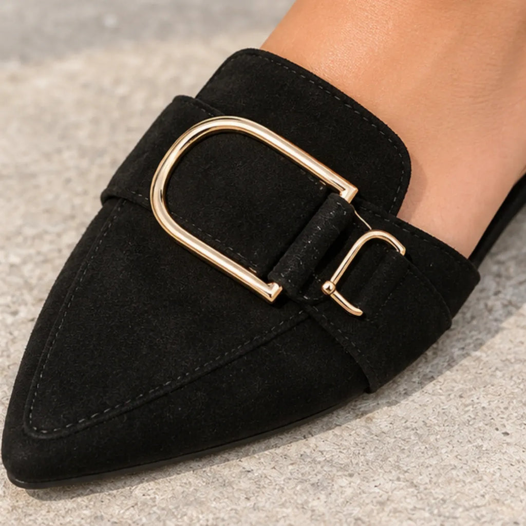 A close-up of a black pointed-toe loafer with a wide strap and a large gold-tone U-shaped buckle, shown worn on a foot against a gray textured floor.