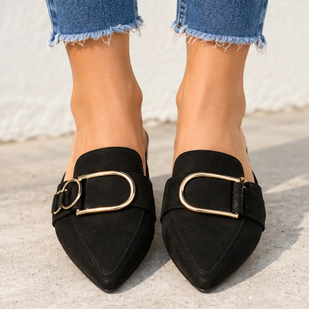 Black pointed-toe slip-on loafers with a wide strap and large gold-tone D-ring buckle, shown on a model wearing frayed blue jeans against a light concrete wall.