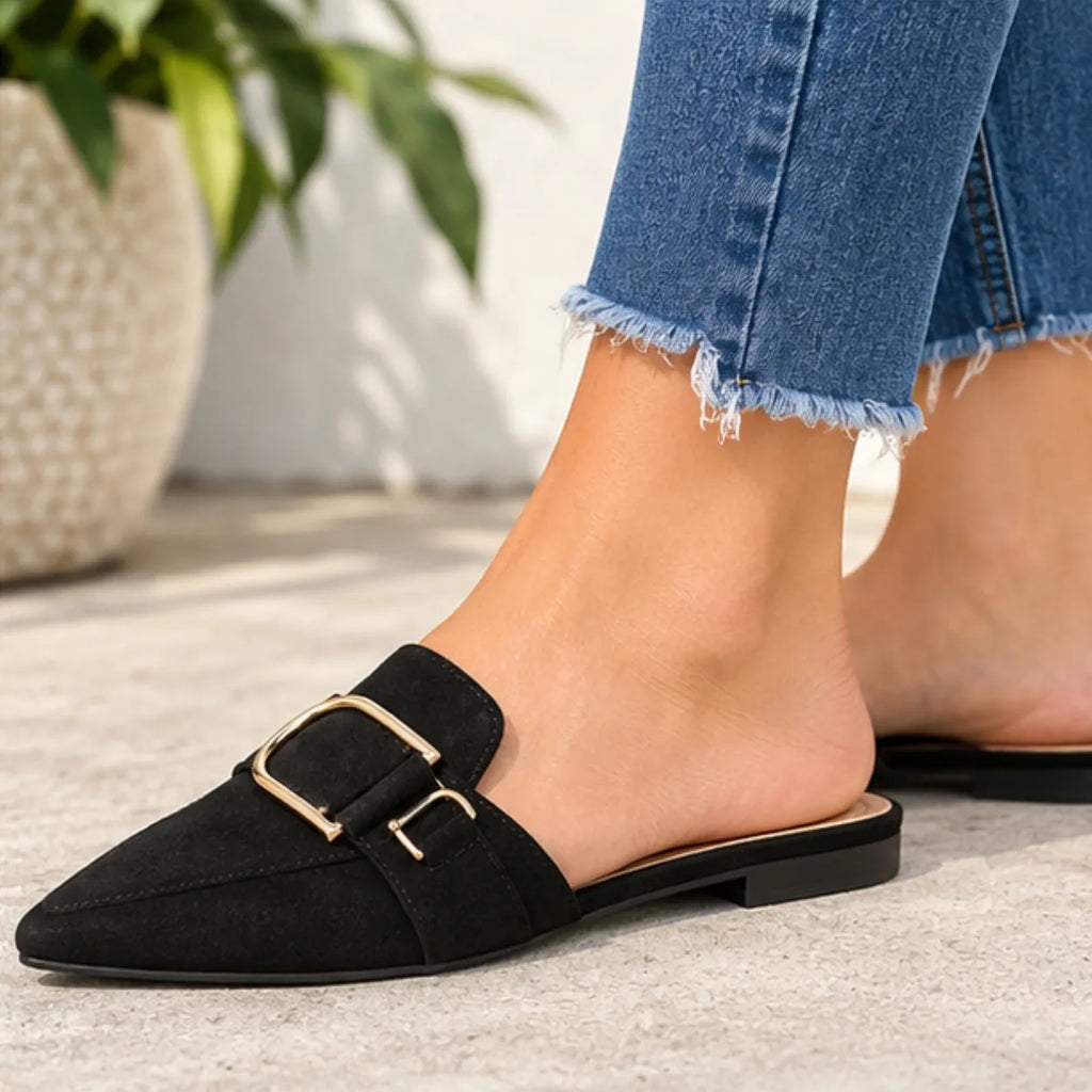 A person wearing black pointed-toe mule flats with a gold-tone double-buckle strap, paired with frayed blue jeans, standing on a light concrete floor.