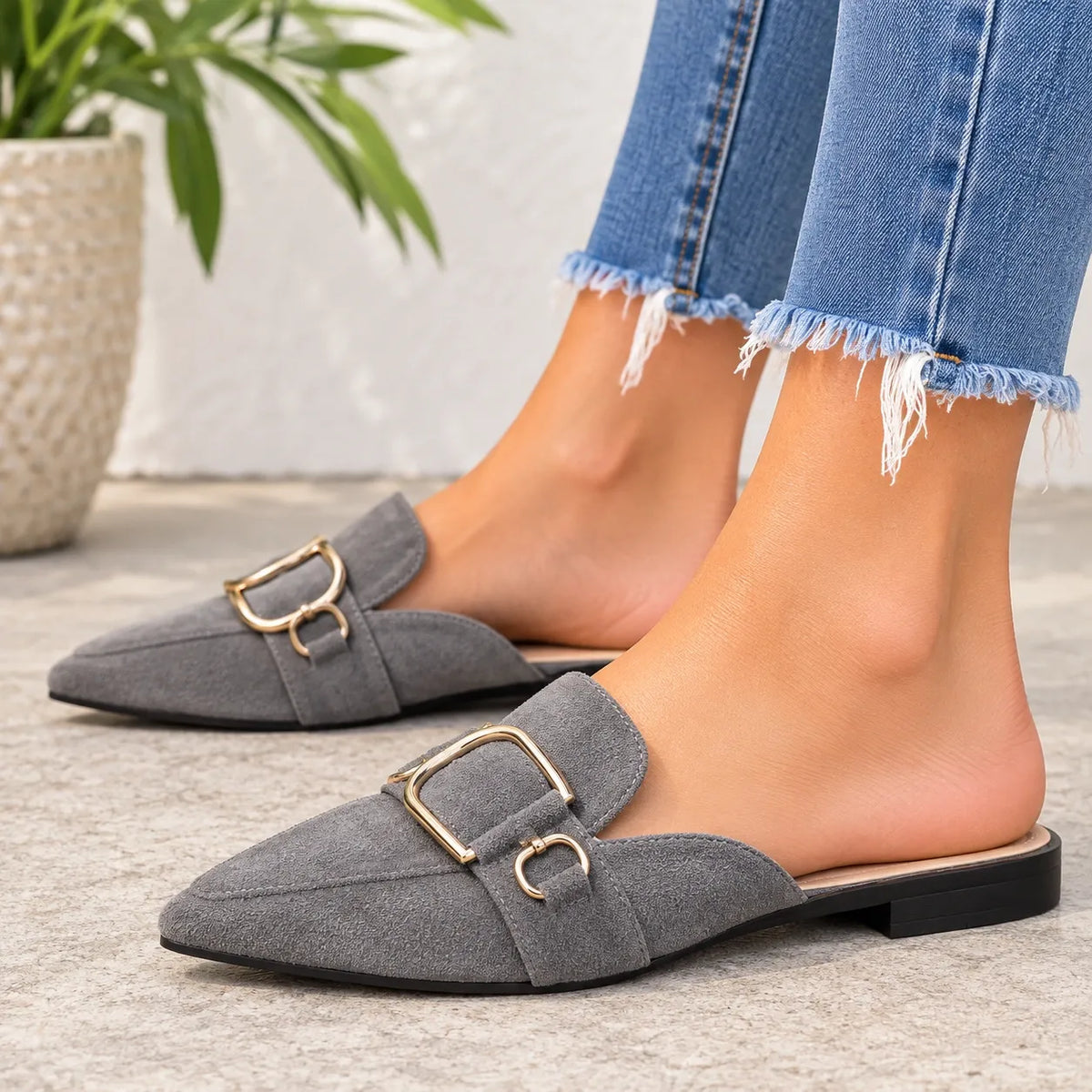 A pair of gray suede mule loafers with gold-tone buckle details, shown on a model's feet wearing frayed blue jeans.