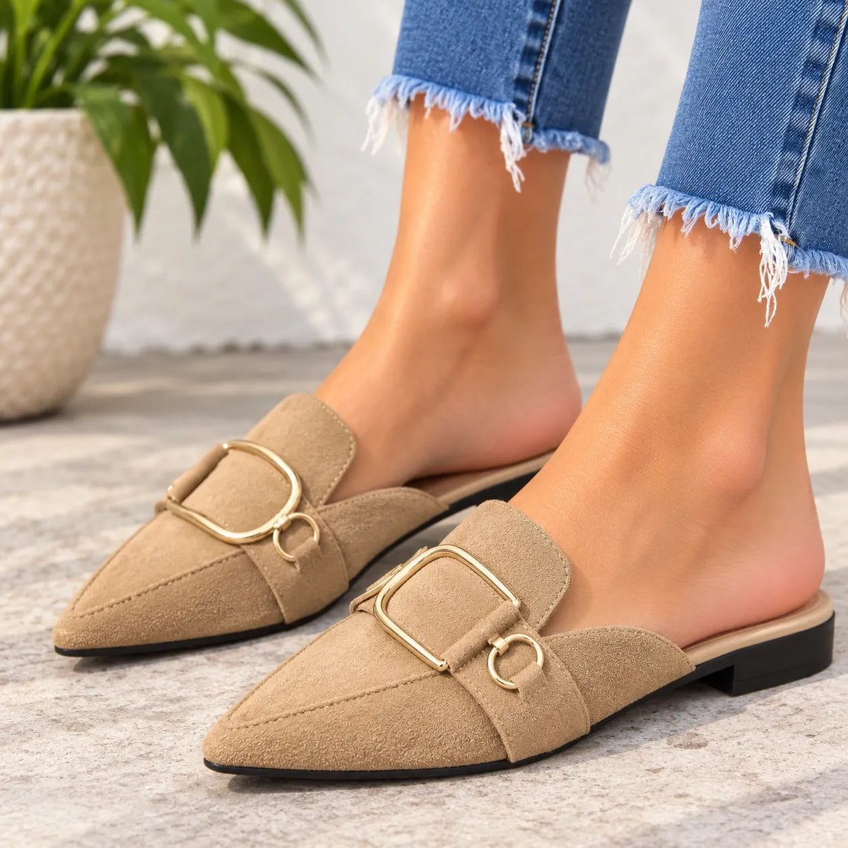 A pair of beige pointed-toe slip-on loafers with gold-tone buckle details, shown on a model's feet wearing frayed blue jeans.