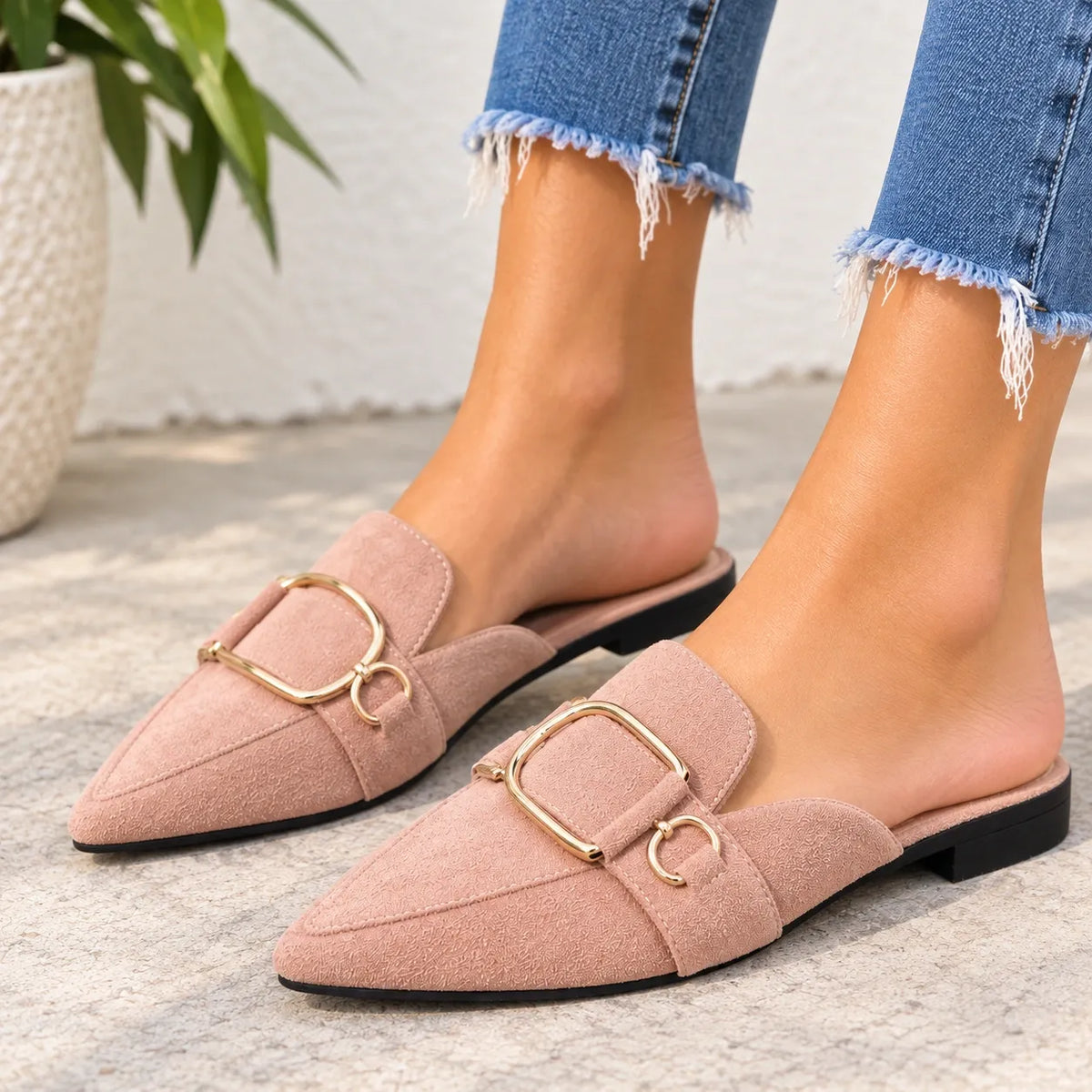 A pair of women's pointed-toe slip-on flats in dusty blush pink suede with gold-tone double-ring hardware, shown on a model wearing frayed blue jeans.