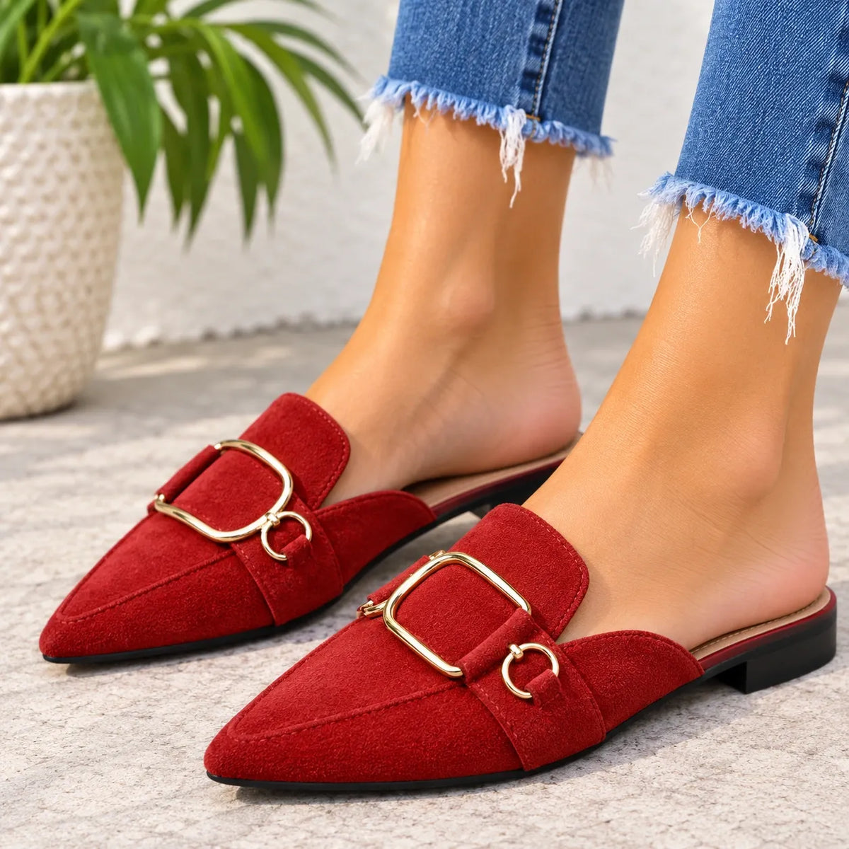 A pair of red suede mule loafers with gold-tone buckle details, shown on a model wearing frayed blue jeans in an outdoor setting.