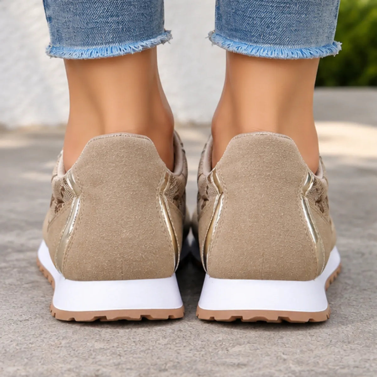 A close-up rear view of a person wearing beige slip-on sneakers with metallic gold side accents and white soles, paired with frayed blue jeans, standing on a concrete surface.
