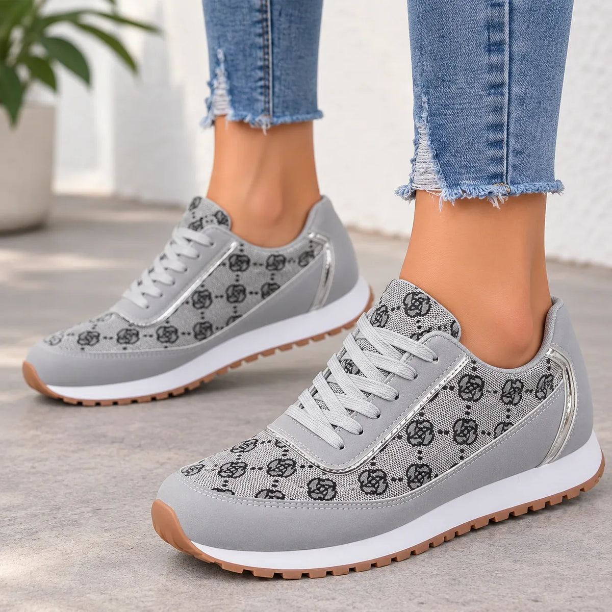 A pair of women's low-top sneakers in light gray suede and mesh with a black monogram pattern, worn with frayed blue jeans. The shoes feature white laces, white midsoles, and tan outsoles, shown on a person's feet.