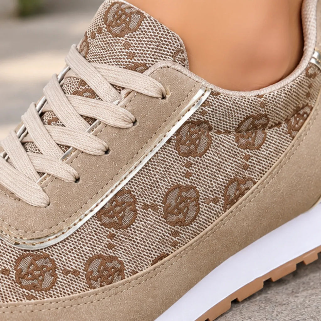 A close-up of a beige low-top sneaker with a woven monogram pattern, beige laces, and a white midsole, shown worn on a foot.