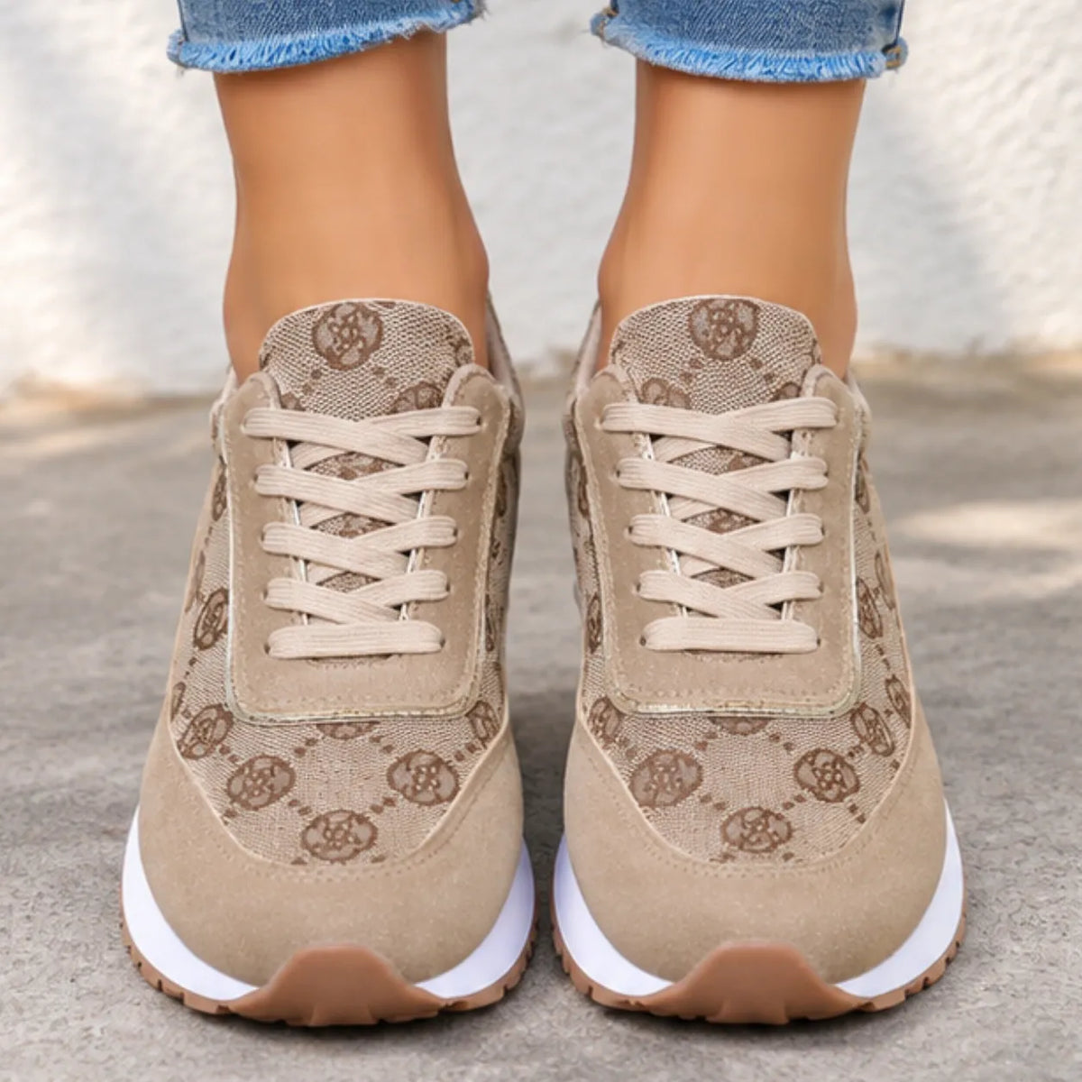A pair of beige low-top sneakers with a brown monogram pattern, worn with frayed blue jeans. The shoes feature beige laces, white midsoles, and brown rubber toe caps, shown from a low-angle shot on a concrete surface.