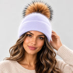 A woman wearing a light lavender knit beanie with a large brown and black faux fur pom-pom, photographed against a plain light gray background.