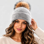 A woman wearing a light gray knit beanie with a large brown and black faux fur pom-pom, paired with a cream knit sweater against a light gray background.