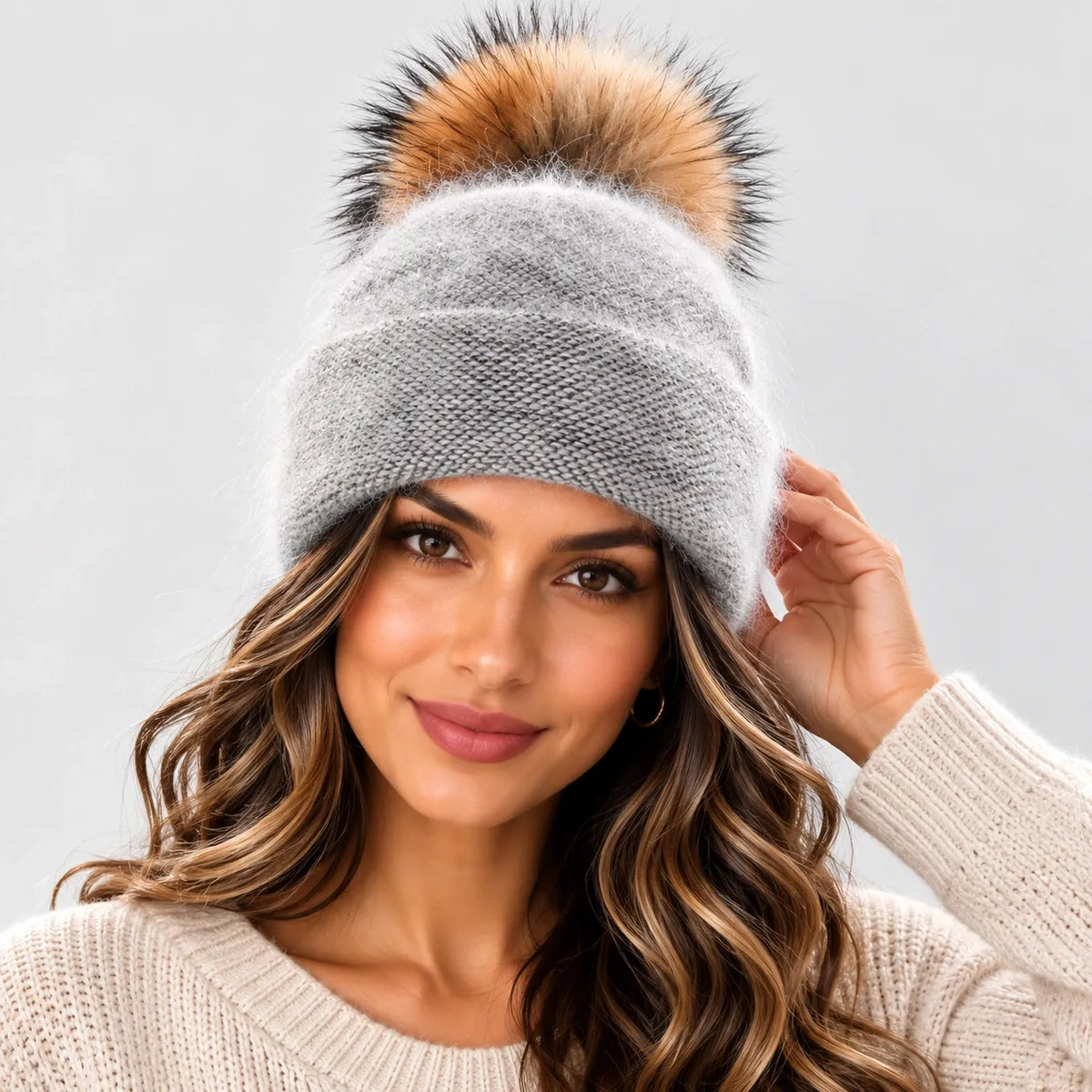 A woman wearing a light gray knit beanie with a large brown and black faux fur pom-pom, paired with a cream knit sweater against a light gray background.