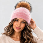 A woman wearing a light pink knit beanie with a large brown and black faux fur pom-pom, paired with a cream-colored knit sweater against a light gray background.