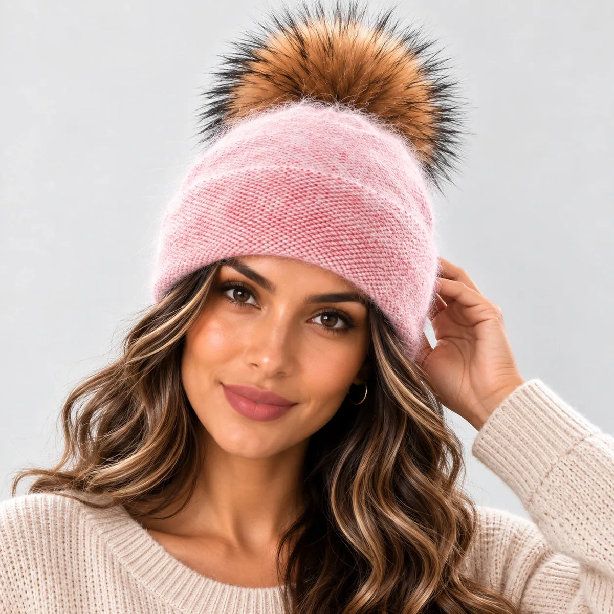 A woman wearing a light pink knit beanie with a large brown and black faux fur pom-pom, paired with a cream-colored knit sweater against a light gray background.