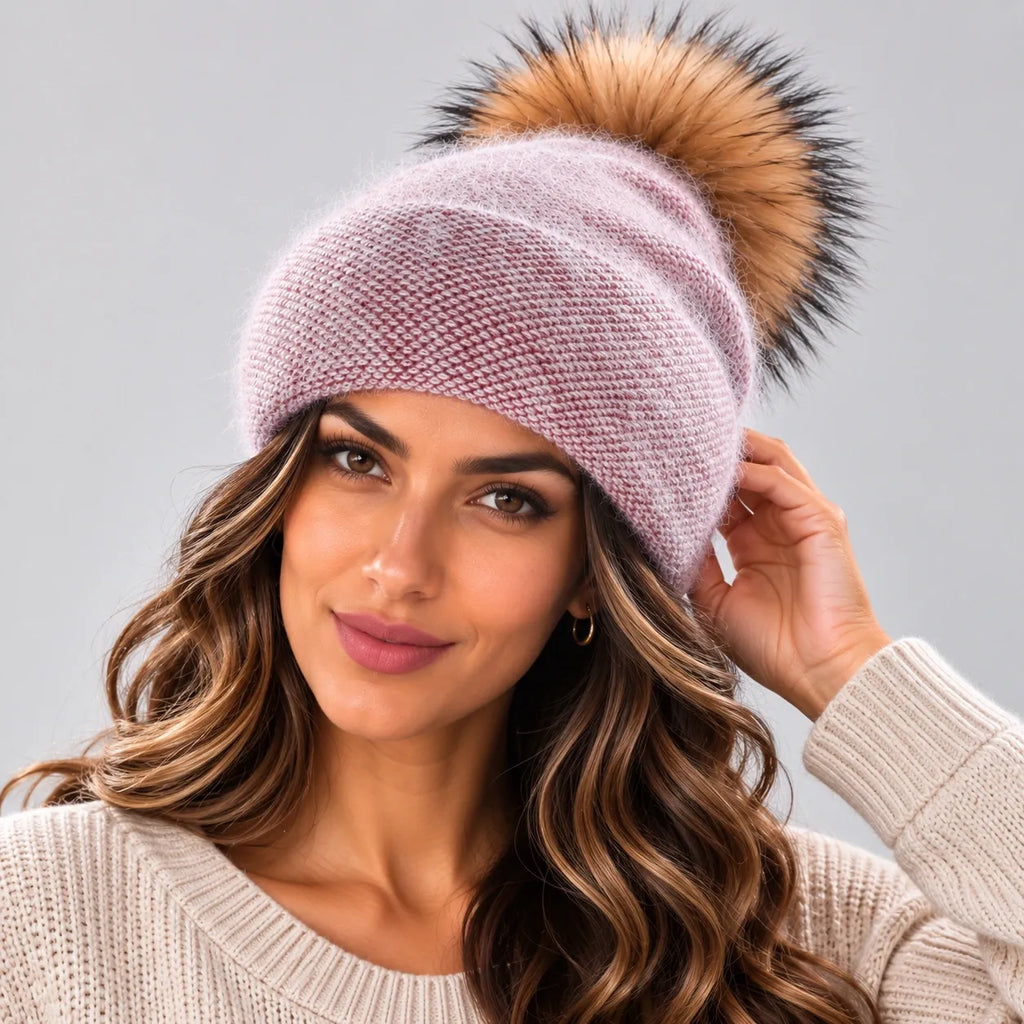 A woman wearing a ribbed knit beanie in dusty pink with a large brown faux fur pom-pom, photographed against a solid gray studio background.