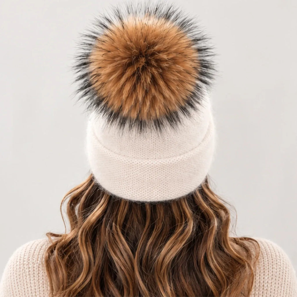A cream-colored knit beanie with a large brown and black faux fur pom-pom, shown from the back on a model with wavy brown hair.