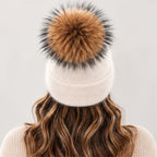 A cream-colored knit beanie with a large brown and black faux fur pom-pom, shown from the back on a model with wavy brown hair.