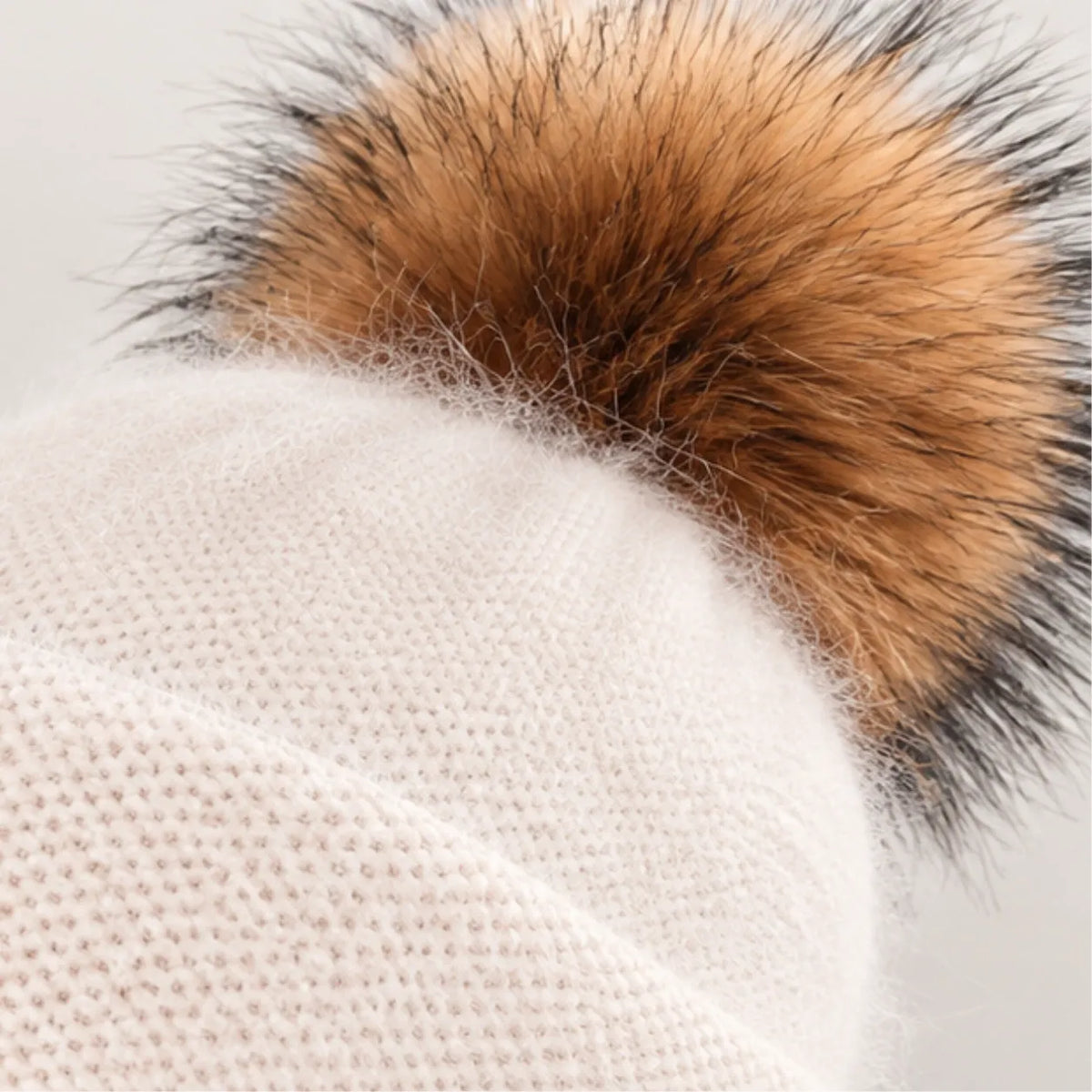 Close-up of a knit beanie hat in a warm off-white or cream color, topped with a large fluffy faux-fur pom pom in tan, brown, and black.