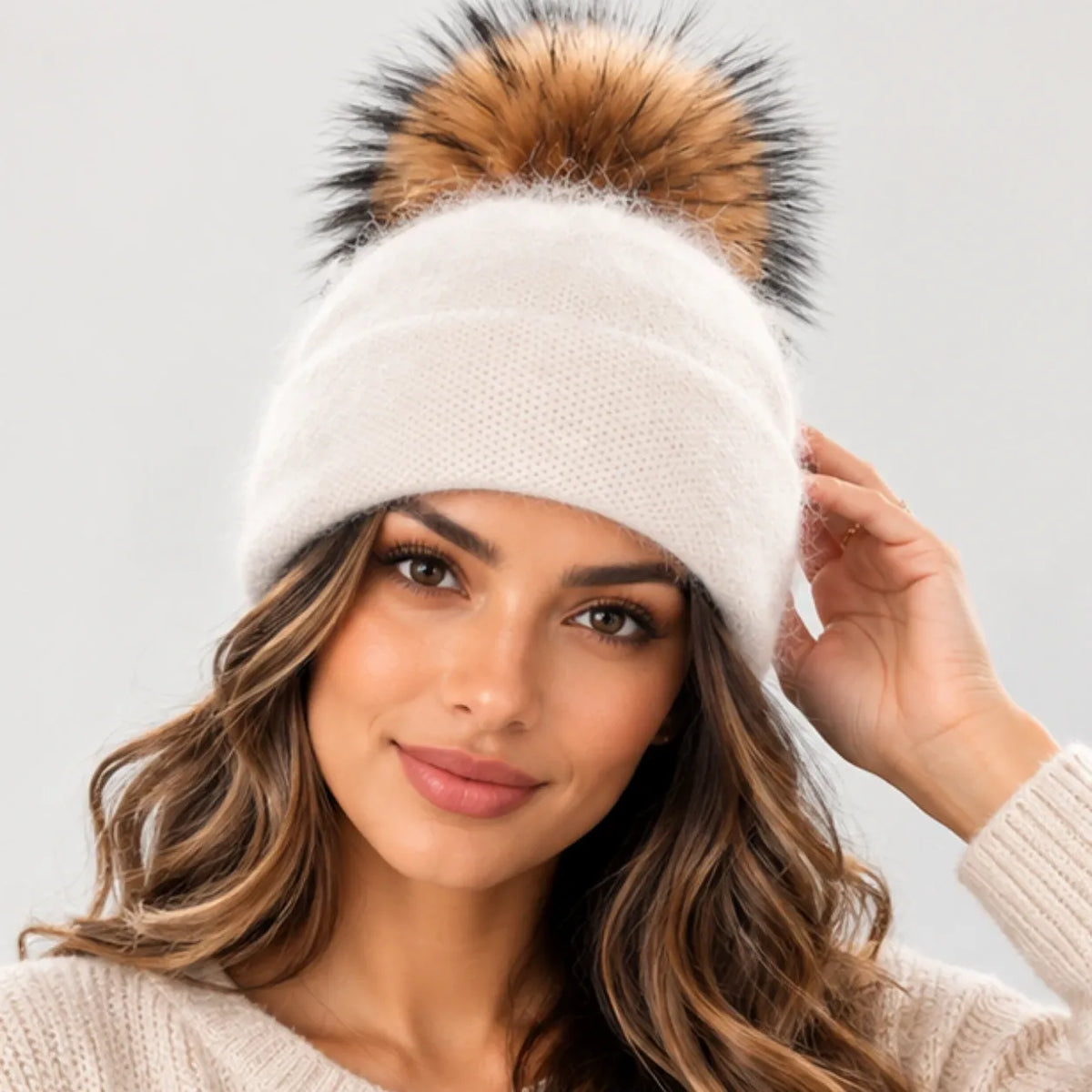 A woman wearing a cream-colored knit beanie with a large brown and black faux fur pom-pom, posing against a light gray background.