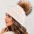 A woman wearing a cream-colored knit beanie with a large brown and black faux fur pom-pom, shown in profile against a light gray background.