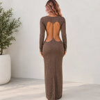 A woman viewed from the back wearing a long-sleeve, floor-length brown dress with a large open back and subtle glitter texture, standing against a light neutral wall next to a potted plant.