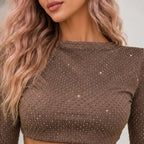A close-up of a woman wearing a brown long-sleeve crop top covered in sparkling rhinestone embellishments. The shot focuses on the textured bodice and neckline, with her wavy blonde hair visible on the left side.