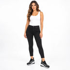 A woman modeling black skinny jeans with a white tank top and black sneakers, standing against a solid white studio background.