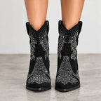 Black suede cowboy-style ankle boots with silver rhinestone western bootleg patterns, shown on a model's feet against a neutral gray background.