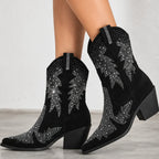 Black suede cowboy boots with silver rhinestone western embroidery, shown on a model's feet against a light gray background.