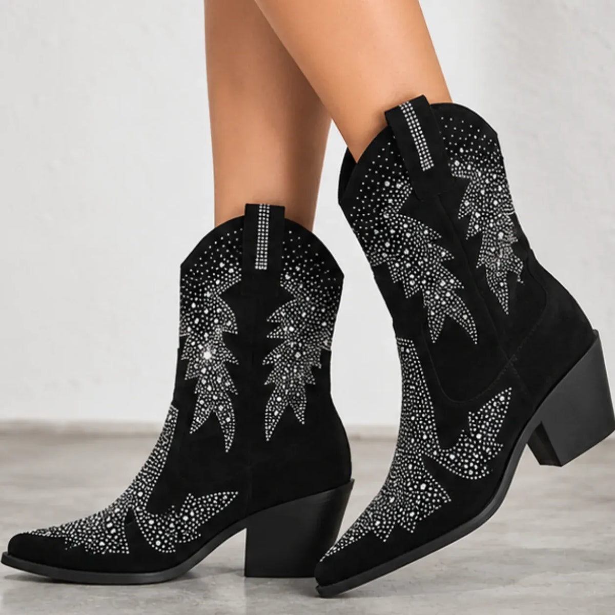 Black suede cowboy boots with silver rhinestone western embroidery, shown on a model's feet against a light gray background.