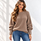 A woman wearing a taupe brown ribbed knit sweater with decorative button details on the sleeves, paired with blue jeans, standing against a white wall with soft shadow patterns.