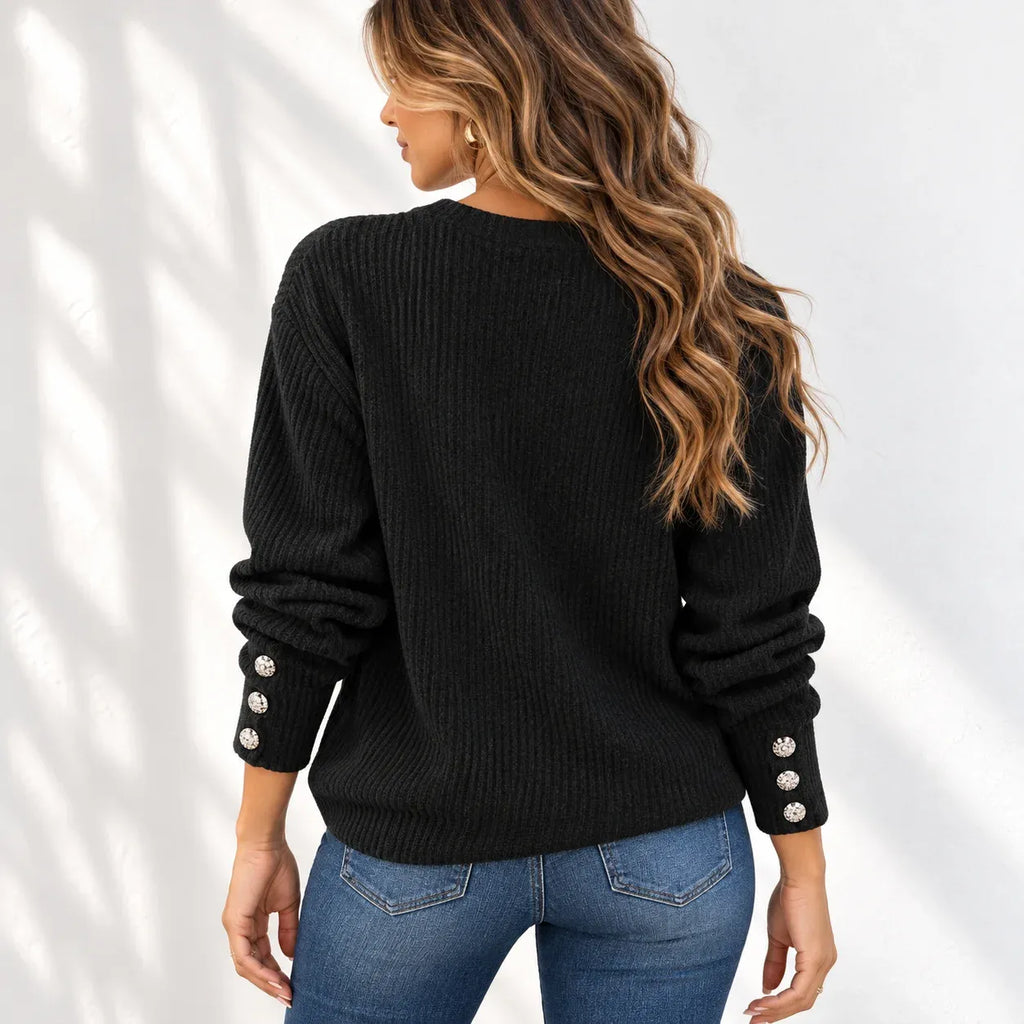 A woman shown from the back wearing a black ribbed knit sweater with decorative silver button details on the cuffs, paired with blue jeans, set against a white wall with soft leaf shadows.