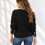 A woman shown from the back wearing a black ribbed knit sweater with decorative silver button details on the cuffs, paired with blue jeans, set against a white wall with soft leaf shadows.