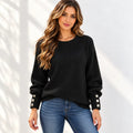 A woman wearing a black ribbed knit sweater with decorative gold-tone buttons on the cuffs, paired with blue jeans, standing against a white wall with soft shadow patterns.
