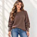 A woman wearing a brown ribbed knit sweater with button details on the cuffs, paired with blue jeans, standing against a white wall with soft shadow patterns.
