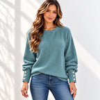 A woman wearing a teal ribbed knit sweater with button details on the sleeves, paired with blue jeans, standing against a white wall with soft shadow patterns.