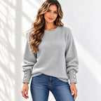 A woman wearing a light gray ribbed knit sweater with decorative silver buttons on the cuffs, paired with blue jeans, standing against a white wall with soft shadow patterns.