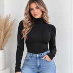 A woman wearing a fitted black ribbed long-sleeve turtleneck top and light blue jeans, standing indoors against a light wall with a vase of dried grass on the left.