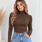 A woman wearing a fitted, long-sleeve ribbed turtleneck top in dark brown, paired with light-wash blue jeans, standing against a light-colored wall.
