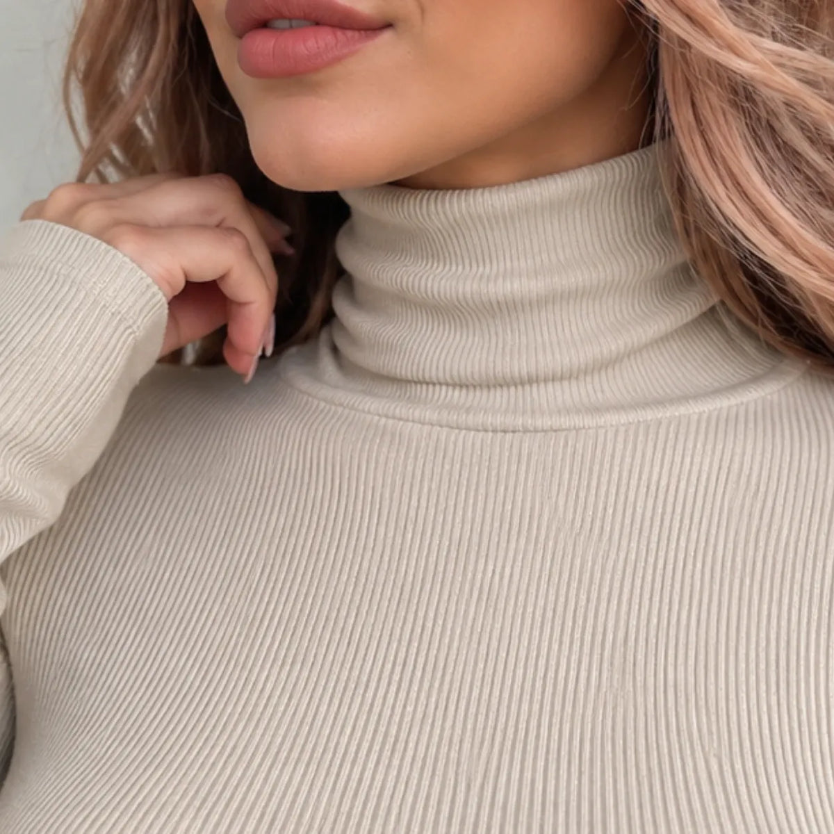 Close-up of a woman wearing a ribbed turtleneck sweater in a light beige color, showing the texture of the knit fabric and her hand resting near her collar.