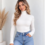 A woman wearing a fitted white ribbed turtleneck long-sleeve top and light blue jeans, standing indoors against a light gray wall with a vase of dried grass on the left.