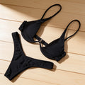 A black two-piece bikini set consisting of a triangle bikini top with adjustable underwire and ruffled details, and matching bikini bottoms, laid flat on a light wood plank background.