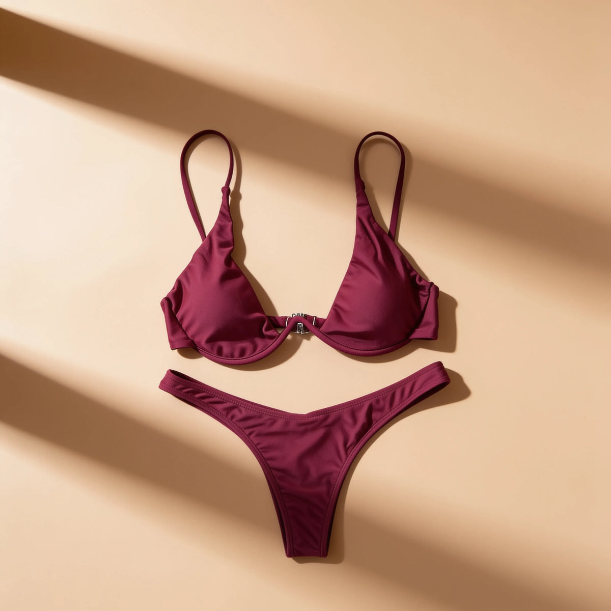 A two-piece bikini set in a deep burgundy or plum color, laid flat on a warm beige background with diagonal shadows.