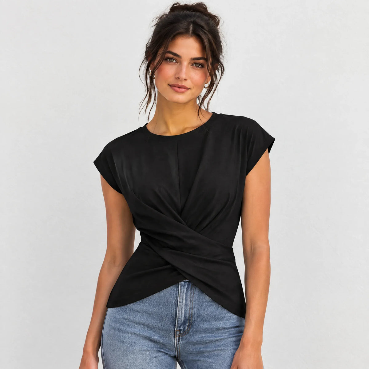 A woman wearing a black short-sleeve top with a twisted wrap-front detail and light blue jeans, photographed against a plain light gray studio background.