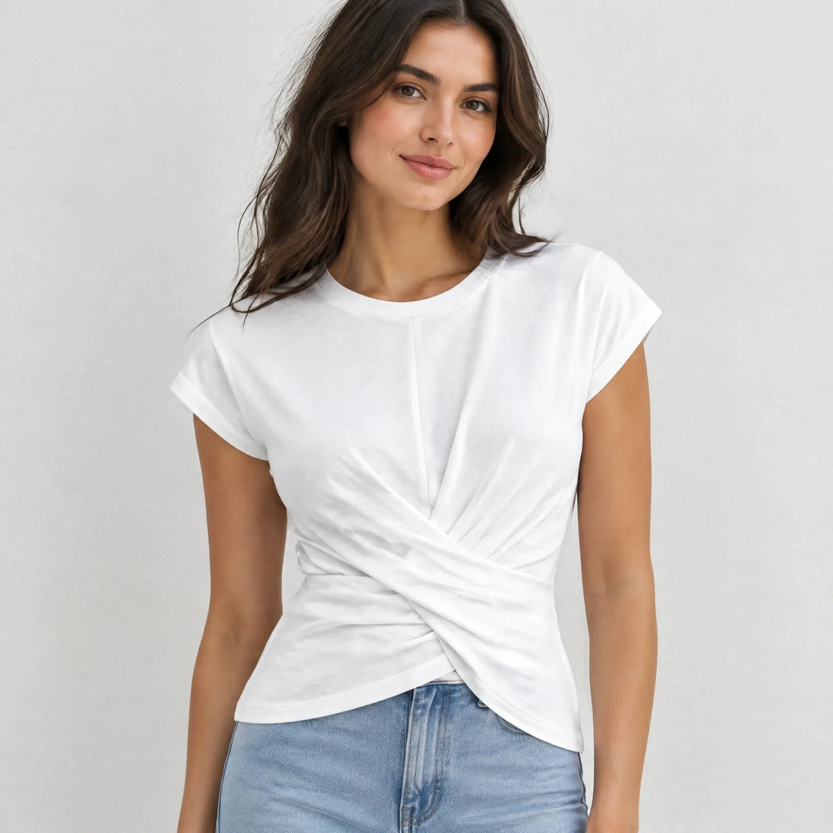 A woman wearing a white short-sleeve t-shirt with a twisted knot detail at the front, paired with light blue jeans, against a light gray background.