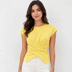 A woman wearing a bright yellow short-sleeve top with a twisted wrap-front detail and white pants, standing against a light gray studio background.