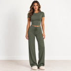 A woman modeling an olive green two-piece lounge set consisting of a short-sleeve crop top and matching wide-leg drawstring pants, standing against a clean white studio background.