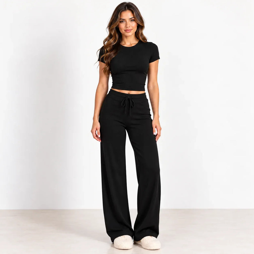 A woman modeling a black short-sleeve crop top and matching black wide-leg drawstring pants against a clean white studio background.
